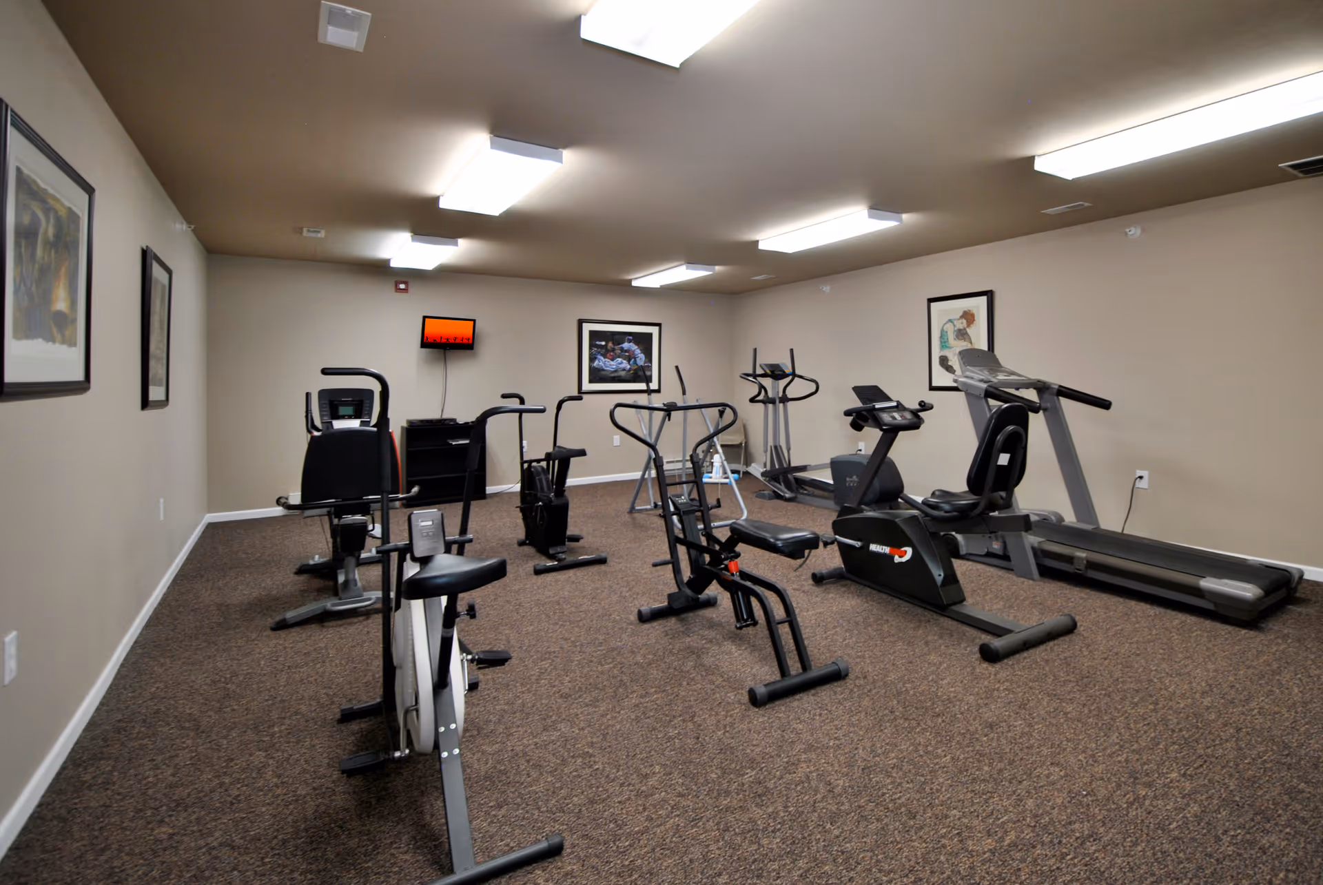 A small fitness room with exercise equipment including stationary bikes, a treadmill, and other workout machines. The room has beige walls, brown carpet, fluorescent ceiling lights, and framed pictures on the walls.