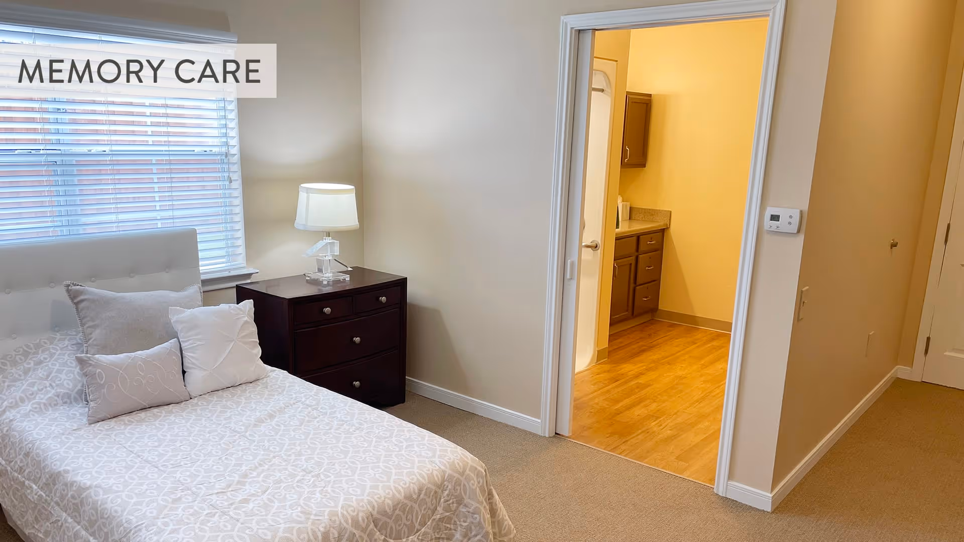A bedroom with a single bed covered in a patterned beige and white bedspread and three pillows. Next to the bed is a dark wooden nightstand with a clear glass lamp. A window with white blinds is behind the bed. An open doorway leads to a room with wooden flooring and cabinets, possibly a bathroom or kitchenette. The words MEMORY CARE are displayed in the upper left corner.