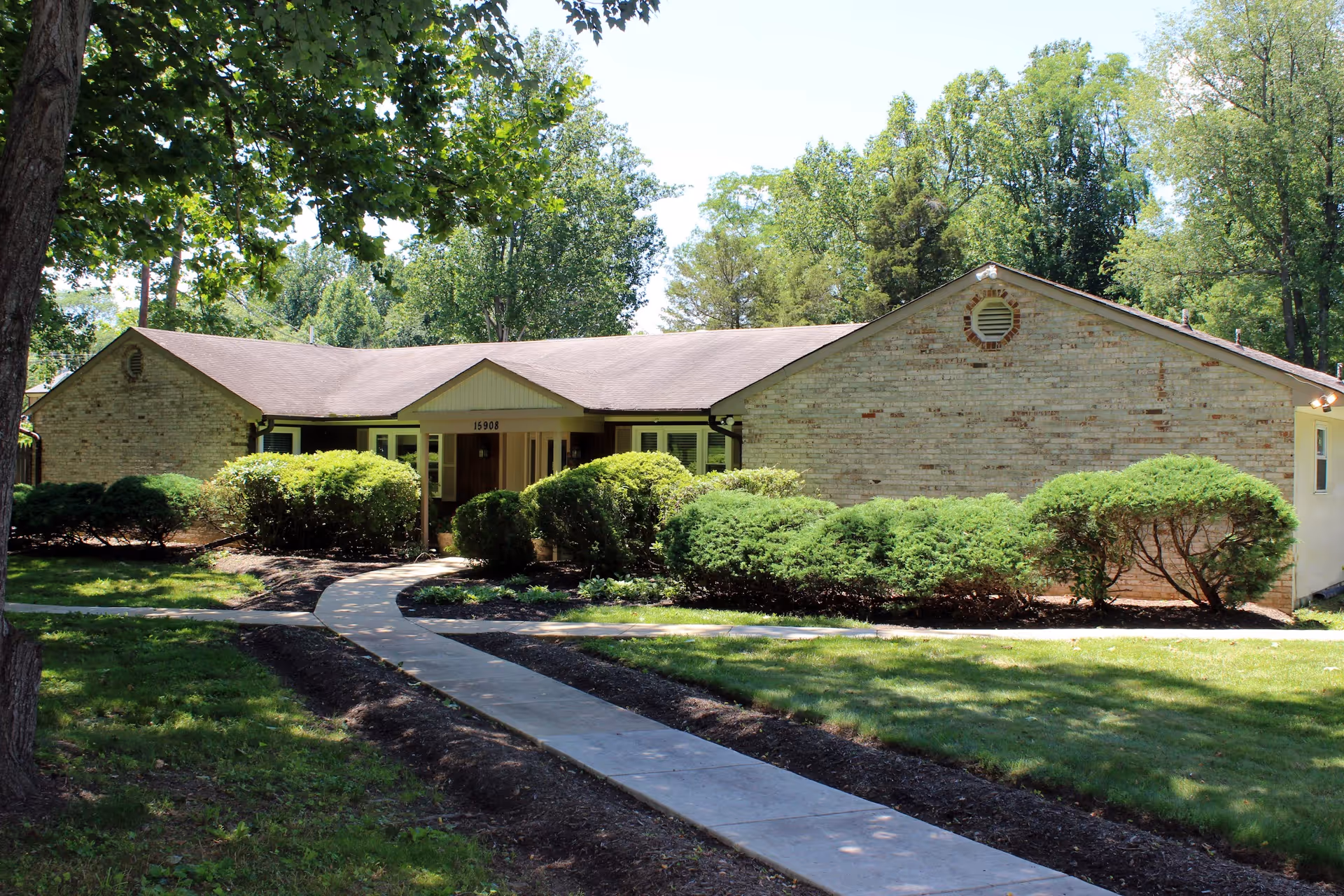 Single-story brick building with a sloped roof surrounded by green bushes and trees. A concrete walkway leads up to the entrance of the building under a small covered porch. The area is well-shaded with sunlight filtering through the trees.