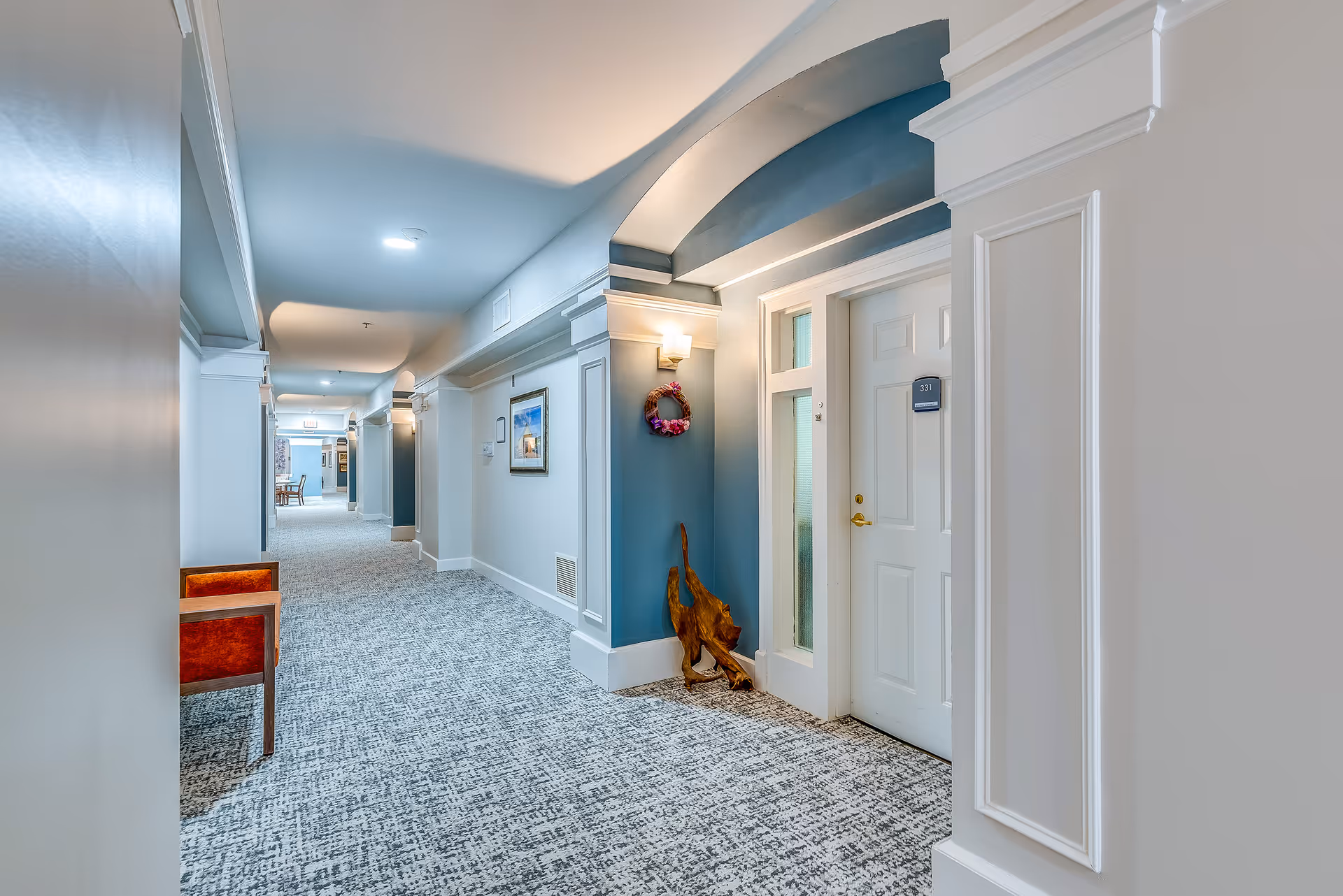 A well-lit, carpeted hallway in a retirement residence with white and blue walls, decorative molding, a white door labeled 331, a small wooden sculpture on the floor, a floral wreath hanging on the wall, and a red cushioned chair along the left side.