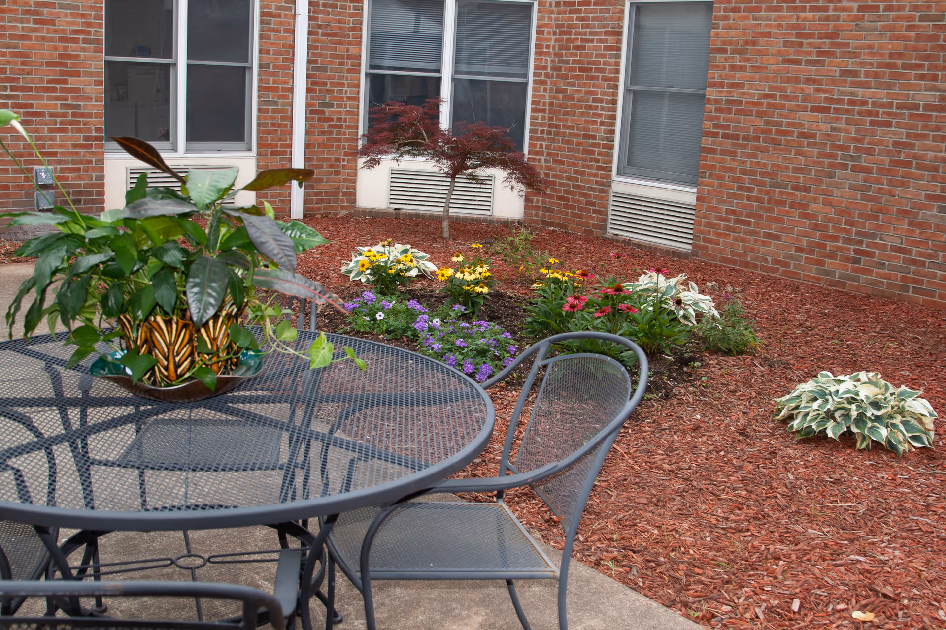 Outdoor patio area with a round metal table and chairs. A decorative plant in a patterned pot is placed on the table. In the background, there is a garden bed with colorful flowers and a small tree, bordered by red mulch. The building's brick exterior and windows are visible behind the garden.