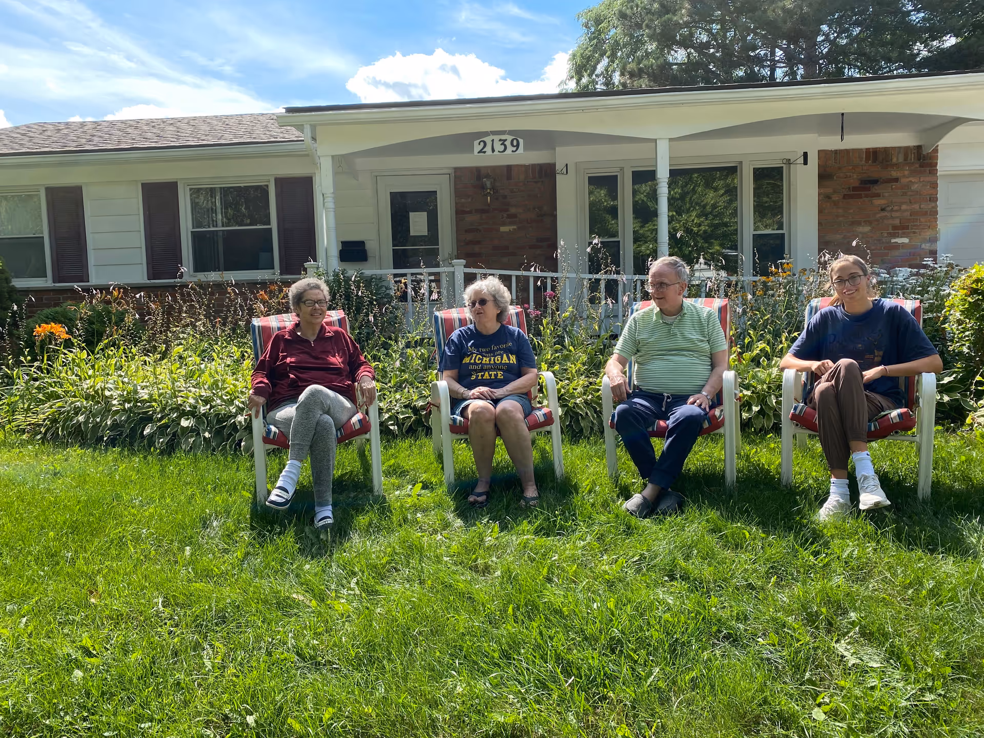 Four people sitting on outdoor chairs on a green lawn in front of a single-story house with a porch and garden. The group includes three older adults and one younger adult, all enjoying a sunny day.