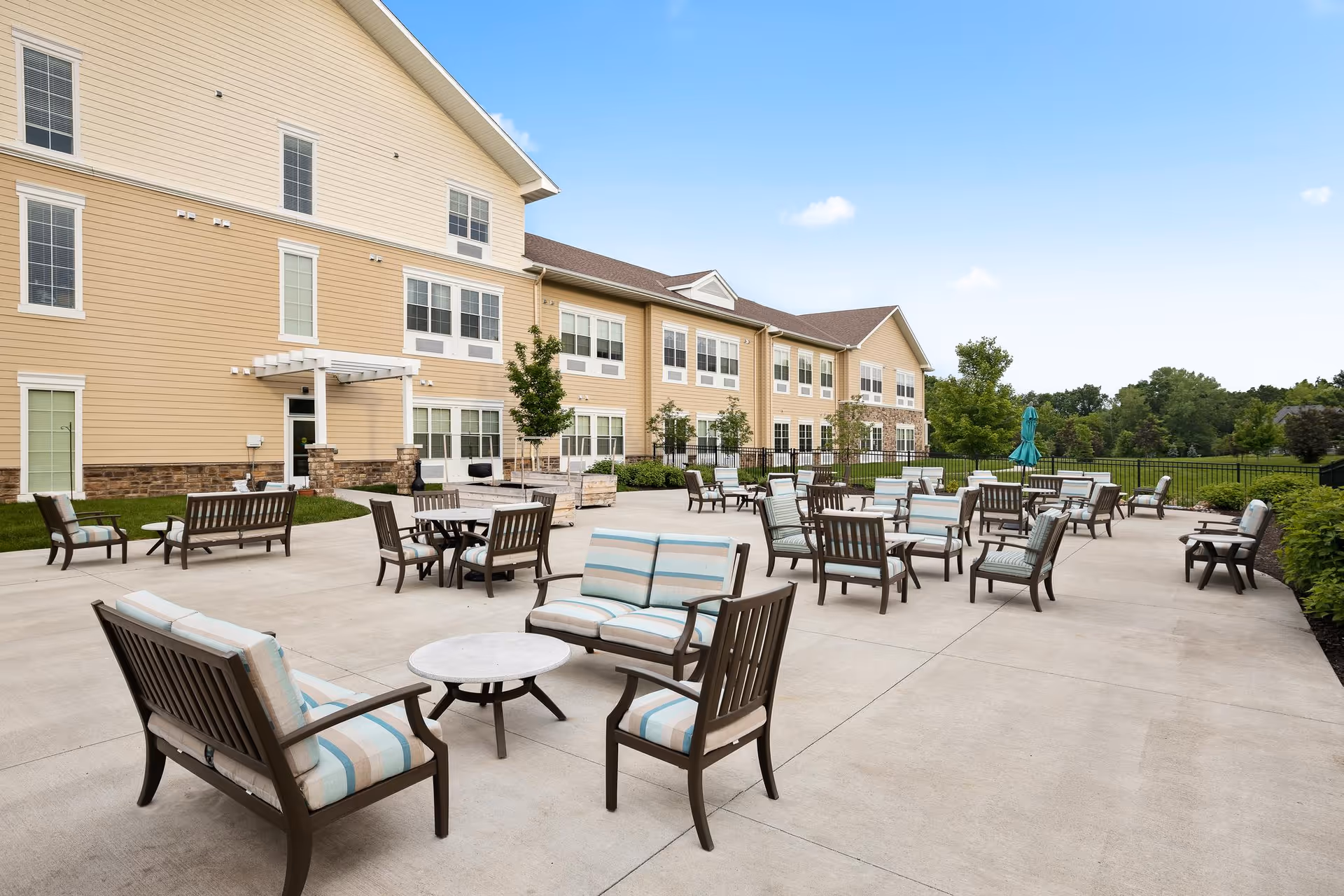 Outdoor patio with multiple cushioned chairs, benches and tables in front of a multi-story beige senior living building under a blue sky.