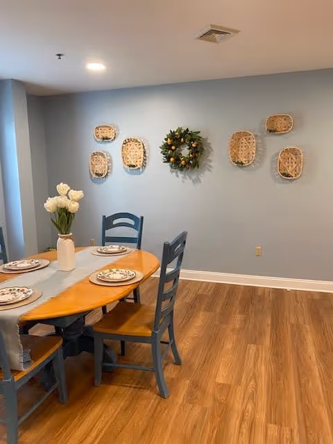 A dining area with a wooden table set for four with floral plates and a vase of white flowers. The chairs are painted blue. The wall behind the table is light blue and decorated with woven baskets and a green wreath with orange accents. The floor is wood with a warm tone.