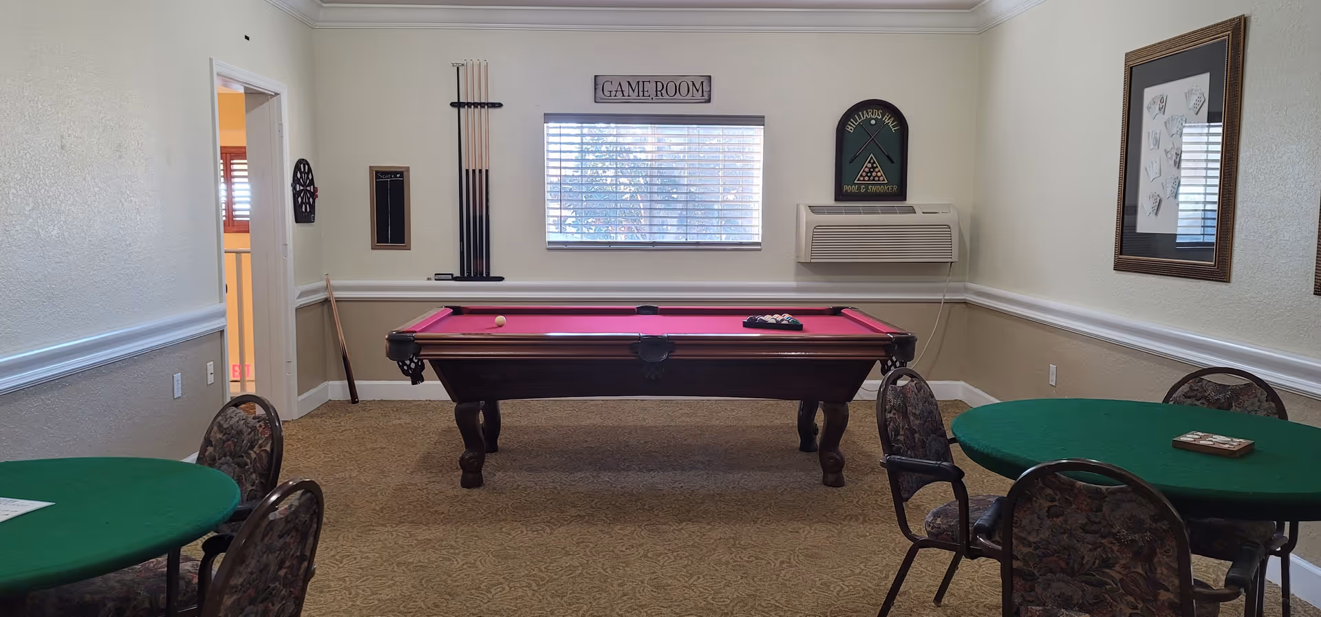 Interior view of a game room with a pool table that has a red felt top in the center. There are two round green card tables with floral patterned chairs around them. On the wall above the pool table is a window with blinds, a sign that says 'GAME ROOM,' and a rack holding pool cues. To the right of the window is an air conditioning unit and a framed sign that reads 'BILLIARDS HALL POOL & SNOOKER.' The walls are painted beige with white trim, and the floor is carpeted.