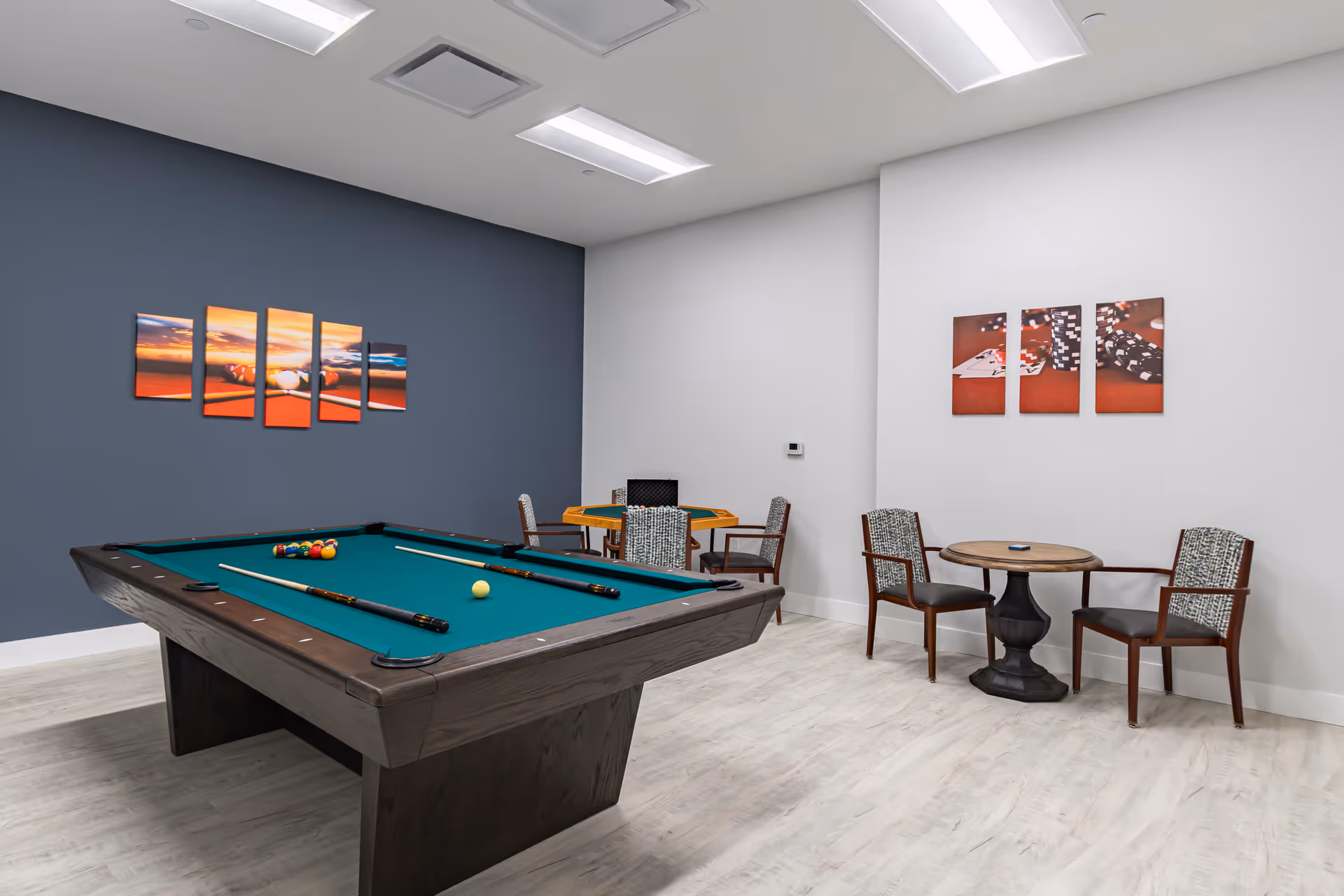 A recreational room with a pool table in the center, two pool cues resting on the table, and billiard balls arranged for a game. The room has light-colored flooring and walls painted white and dark blue. There are two small round tables with chairs around them, and wall art featuring images of billiard balls and poker chips.