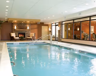 Indoor swimming pool with clear blue water, surrounded by a white tiled deck. In the background, there is a cozy seating area with two chairs and a stone fireplace. Large windows on the right side reveal an adjacent room with exercise equipment.