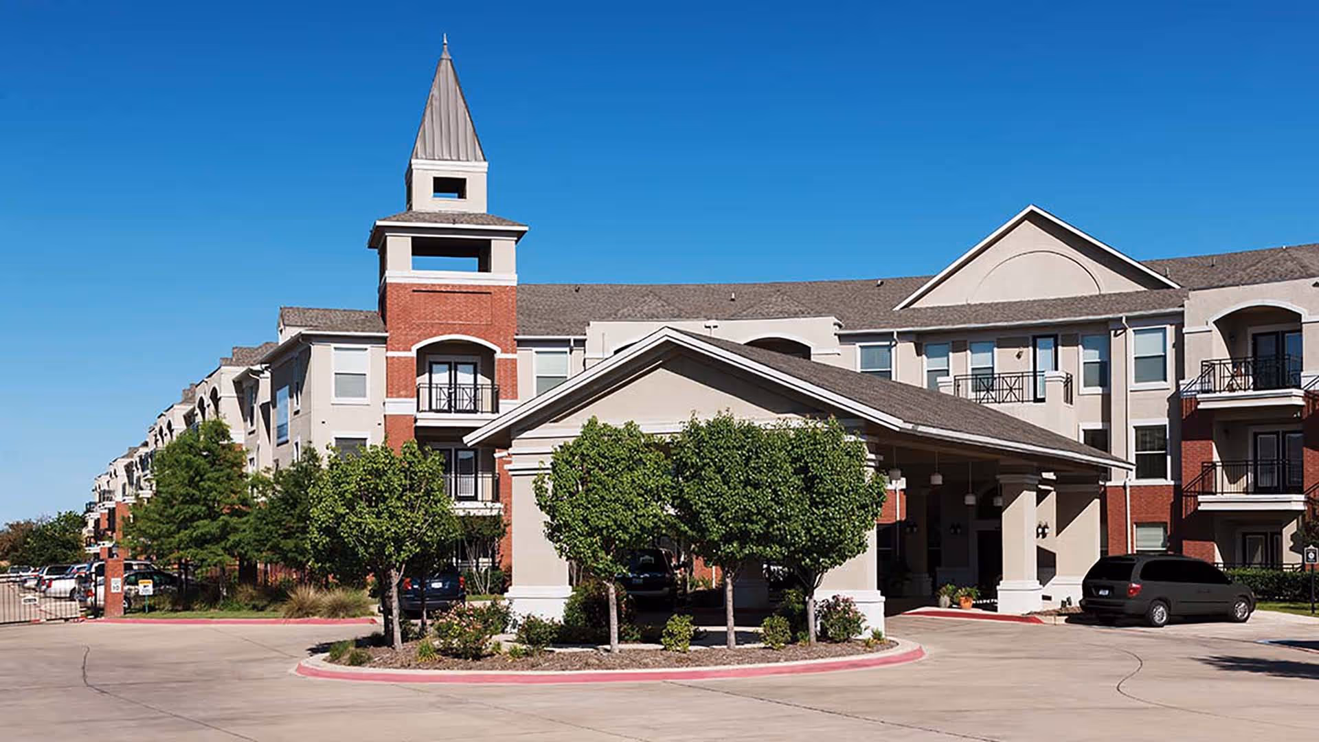 Exterior view of a senior living facility building with a covered entrance, multiple floors, balconies, and a tower-like structure. There are trees and shrubs in front, a clear blue sky, and a parking area with cars.