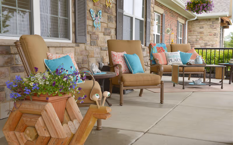 A cozy outdoor patio area with cushioned chairs adorned with colorful pillows, a small table, and decorative flower pots including a wooden snail-shaped planter filled with purple and white flowers. The patio is adjacent to a stone and brick wall with butterfly decorations and hanging flower baskets.