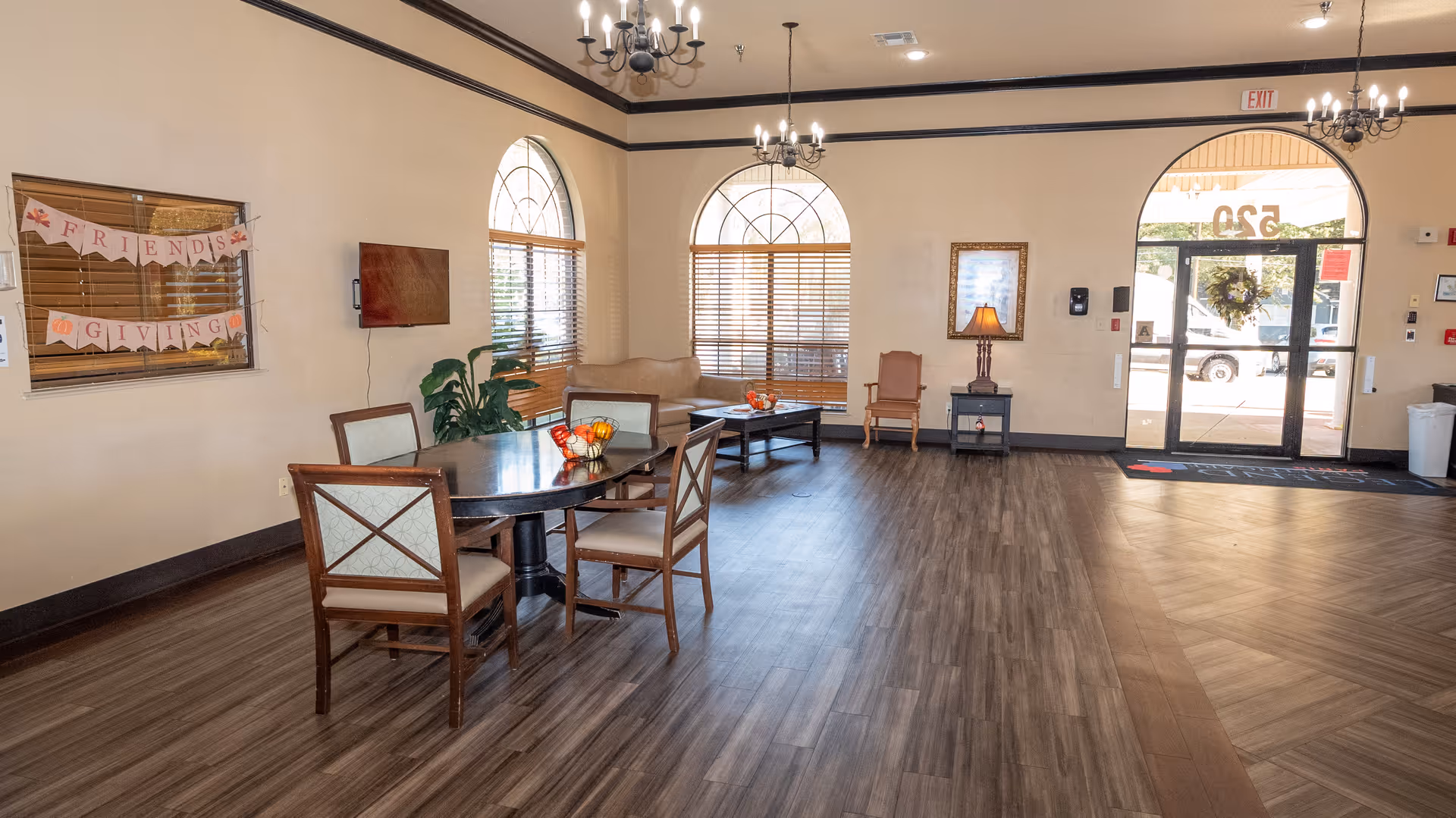 Spacious senior living facility common area with a dining table and chairs, seating area, chandeliers and glass entrance doors.