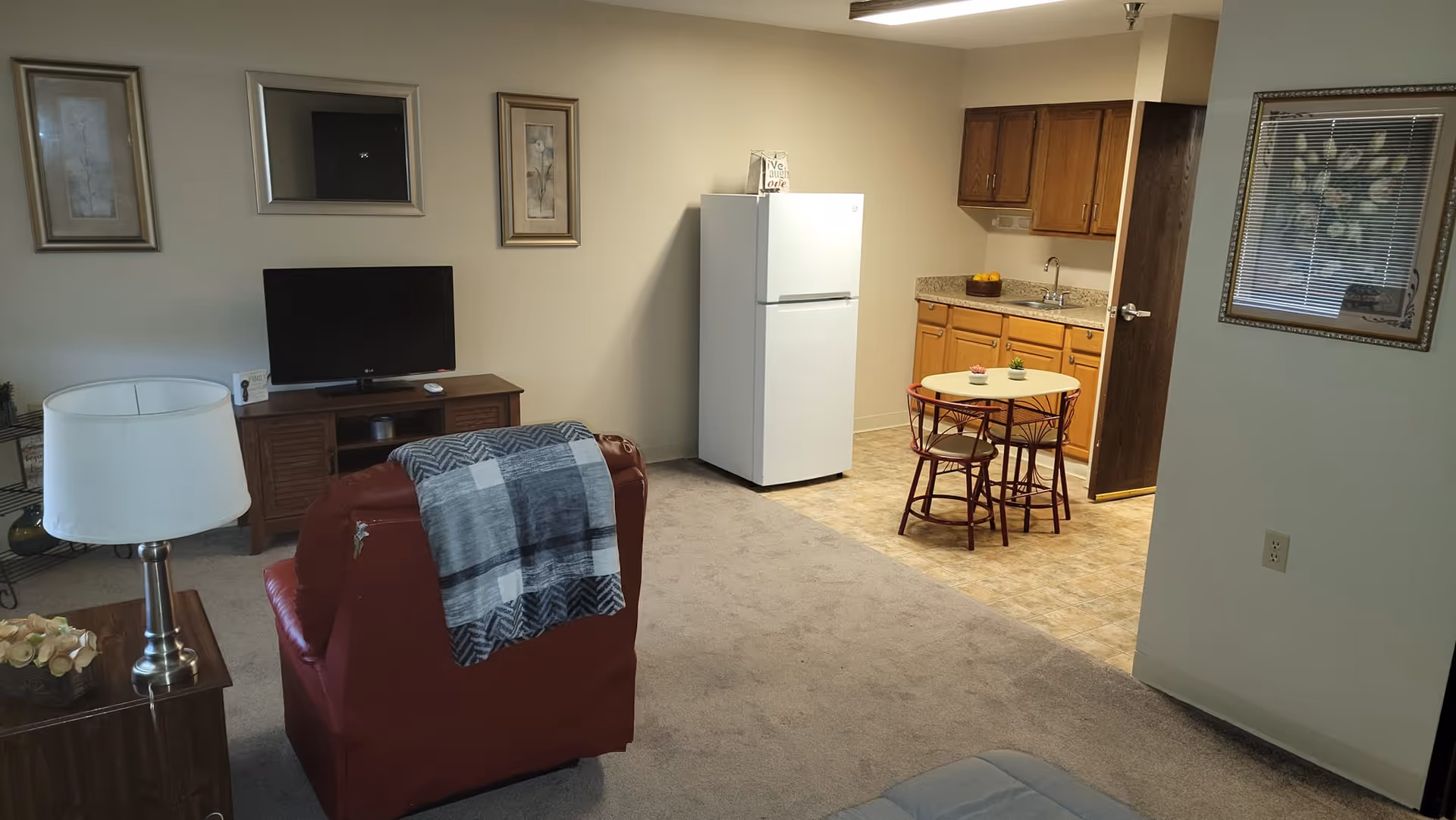 Interior view of a senior living community apartment featuring a red armchair with a gray and white blanket draped over it, a wooden TV stand with a flat-screen TV, a white refrigerator, a small round dining table with two chairs, wooden kitchen cabinets, a countertop with a sink, and framed artwork on the walls.