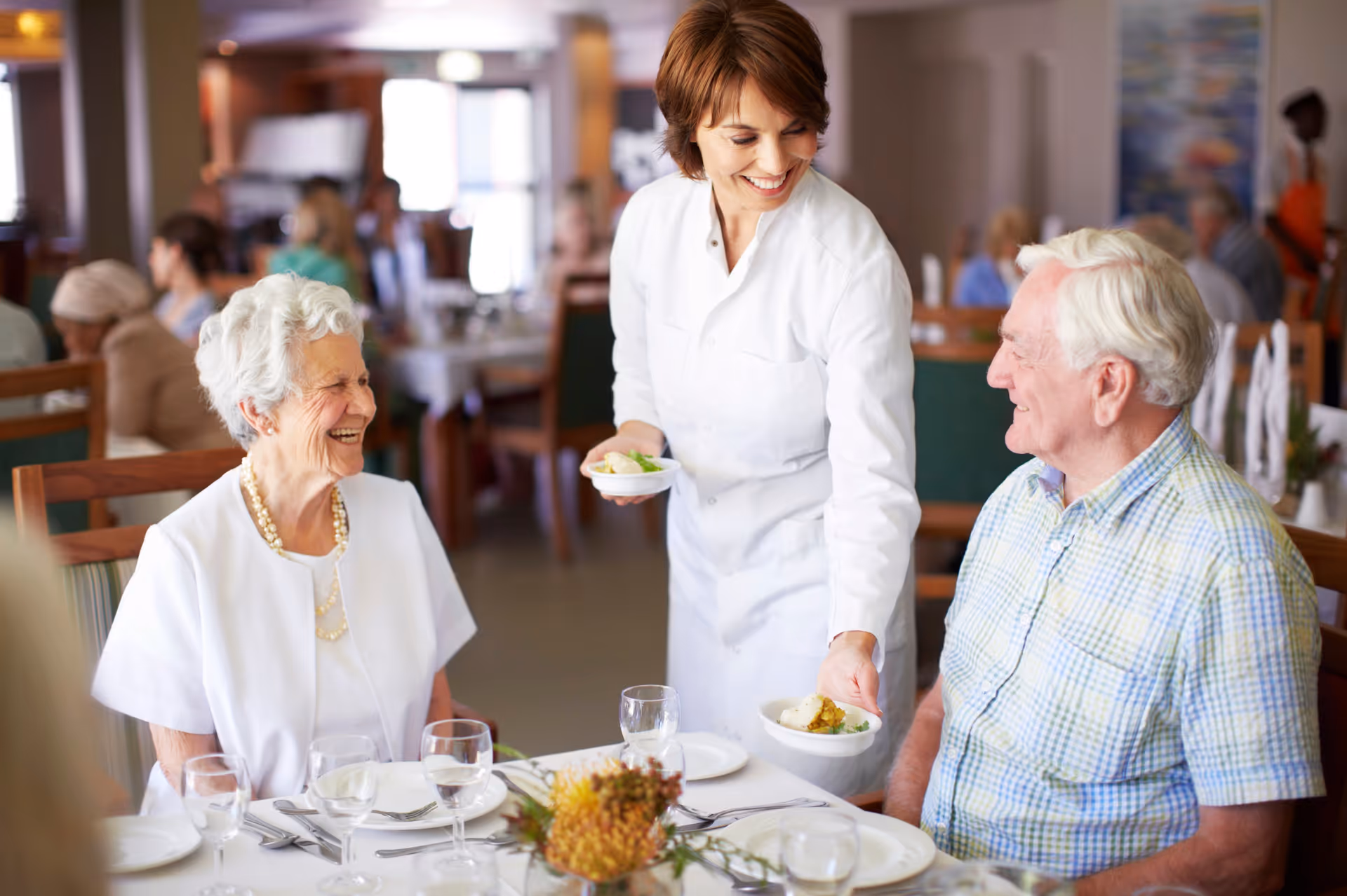 A smiling female server in a white uniform is serving food to an elderly man and woman seated at a dining table in a busy dining room. The elderly woman is wearing a white blouse and a pearl necklace, and the elderly man is wearing a light blue plaid shirt. The table is set with plates, glasses, and silverware, and there are other people dining in the background.