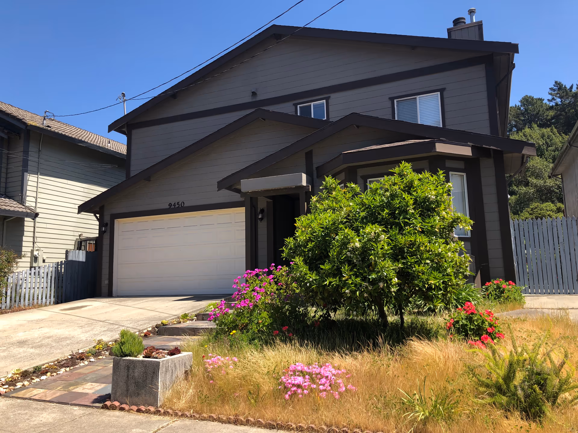 Exterior view of a two-story gray house with a white garage door and the number 9450 above it. The front yard has a small tree, various flowering plants, and a patch of dry grass. The sky is clear and blue.