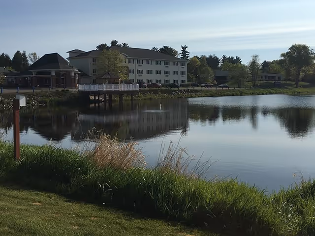 A three-story white senior living building beside a pond, its reflection visible with grassy foreground and a small lakeside deck.