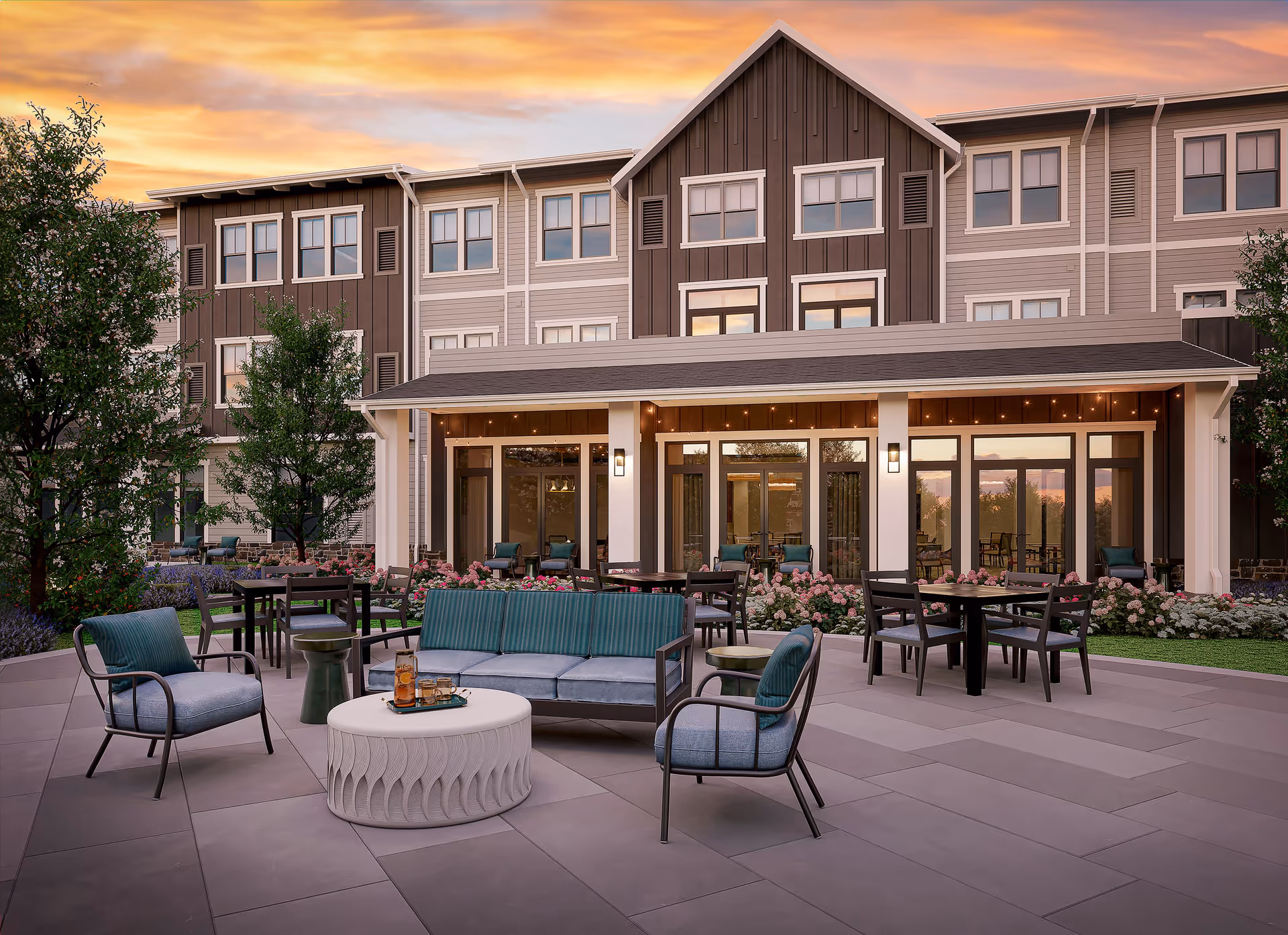 Outdoor patio area at Benchmark at Mount Pleasant with cushioned chairs, a sofa, round tables, and dining tables surrounded by flowers and greenery, set against a multi-story building under a colorful sunset sky.