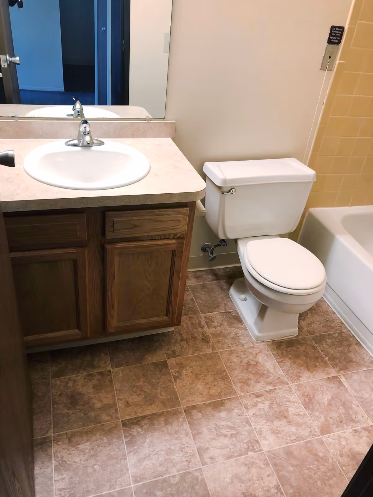 A bathroom with a white sink set in a beige countertop with wooden cabinets below, a white toilet, and a bathtub with yellow tiled walls. The floor is covered with brownish tiles.