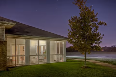 Evening view of a single-story building with a porch illuminated by lights, a tree in the foreground, and a body of water in the background under a dark sky.