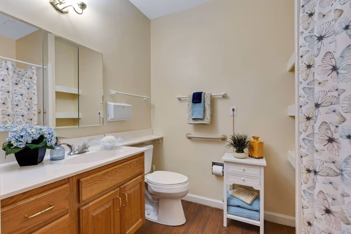 A clean bathroom with a wooden vanity cabinet, white countertop, and a sink with a faucet. There is a mirror above the sink and a small potted plant with blue flowers on the countertop. A toilet is positioned next to the vanity. On the right side, there is a white small cabinet with folded towels and decorative items on top. A towel rack with folded towels is mounted on the wall above a grab bar. A shower curtain with a butterfly pattern is partially visible on the right side.