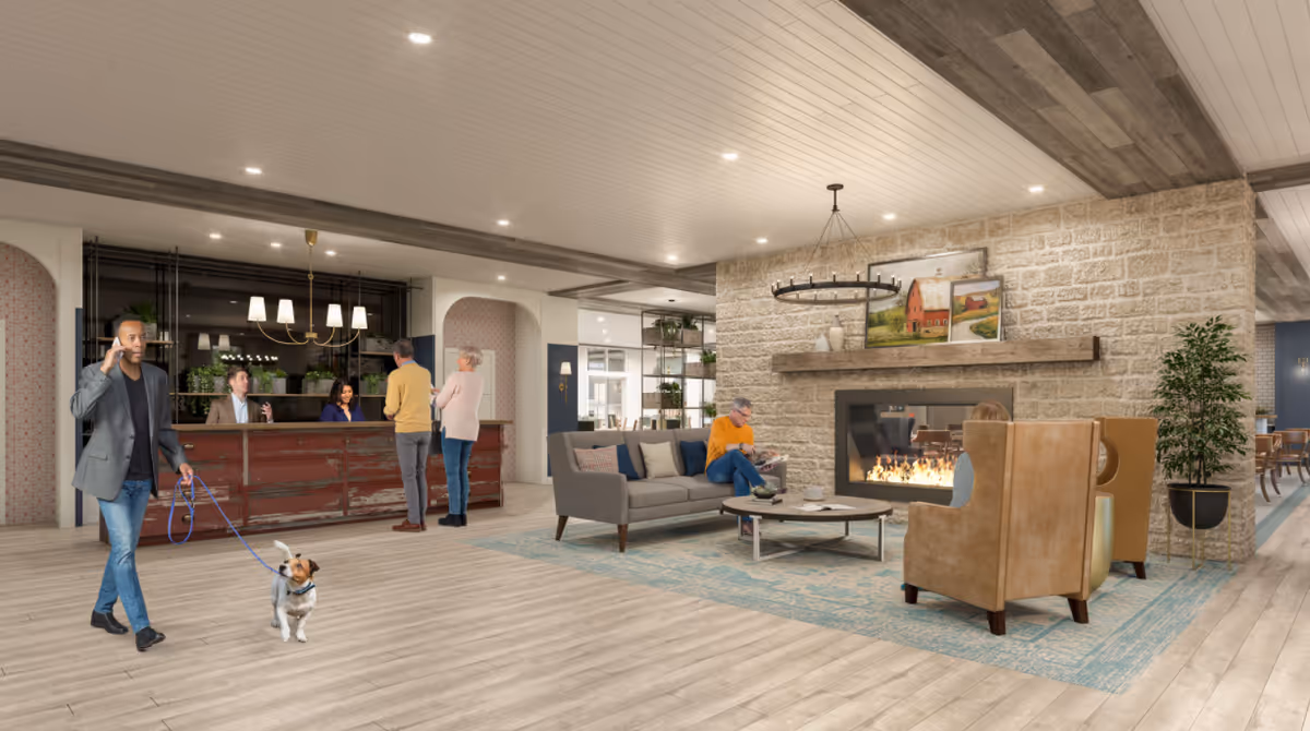 A spacious senior living facility lobby with a man walking a dog on a leash while talking on the phone. In the background, a reception desk with two staff members and two visitors is visible. To the right, there is a cozy seating area with a gray sofa, two tan armchairs, a coffee table, and a stone fireplace with a fire burning. The room is decorated with paintings, plants, and modern lighting fixtures.