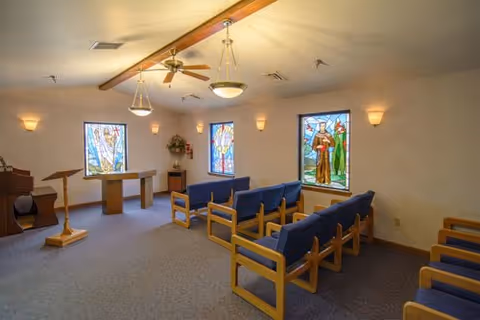 Interior of a small chapel or meditation room with wooden pews featuring blue cushions, stained glass windows depicting religious figures, a wooden lectern, and a piano in the corner. The room is softly lit with wall sconces and ceiling lights.