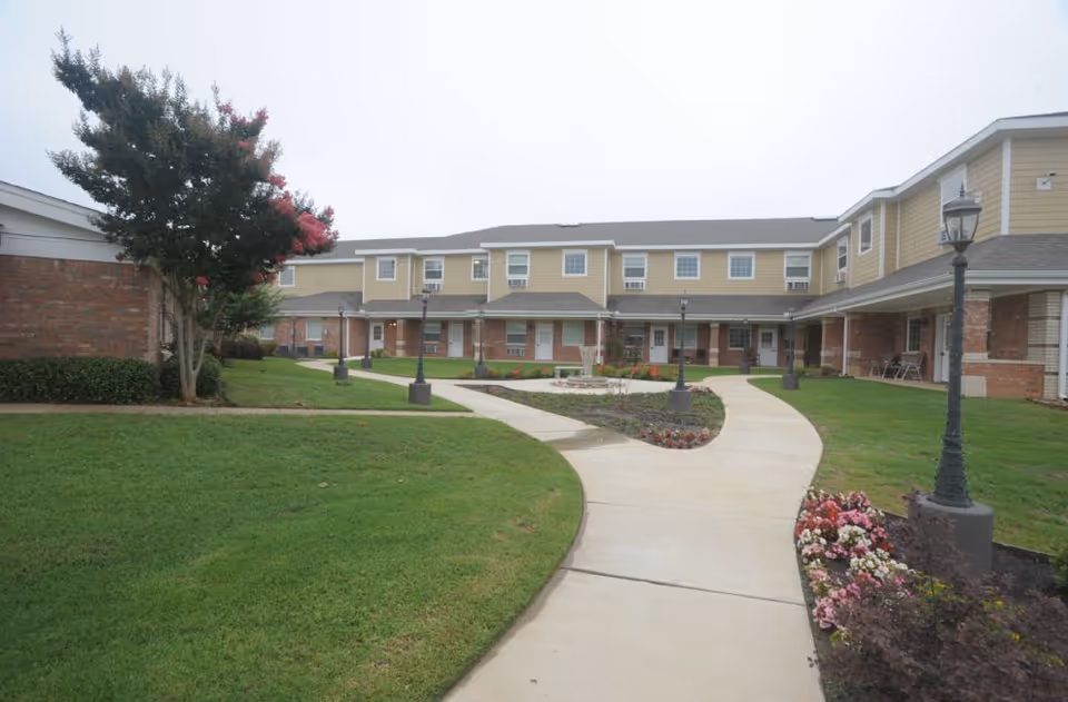 Outdoor courtyard area of Oak Wood Place Assisted Living with a curved concrete walkway, green grass, flower beds, lamp posts, and a two-story building with beige siding and brick accents in the background.