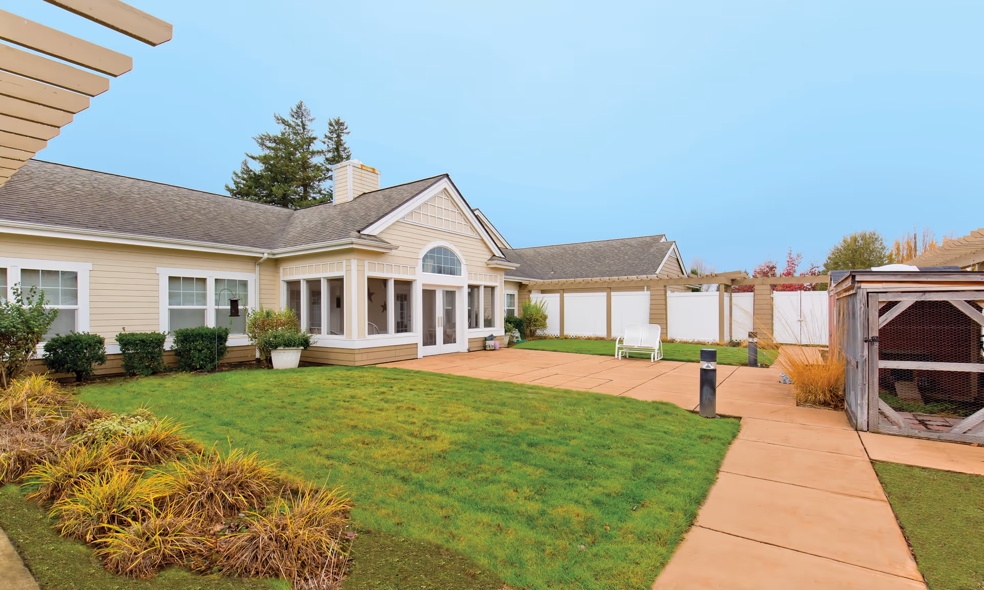 Courtyard of a single-story senior living building with a grassy lawn, paved walkways, patio chairs, a pergola, and a small wooden shed.