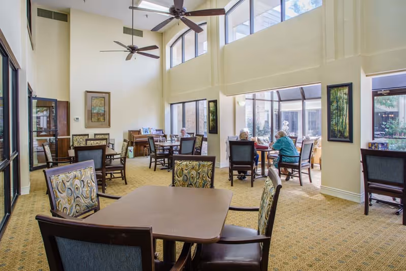 Bright, high-ceiling common dining area with multiple tables and chairs and people seated near large windows.