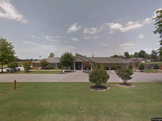 Front exterior view of a single-story brick building with a covered entrance, surrounded by a driveway, green lawn, and small trees under a partly cloudy sky.