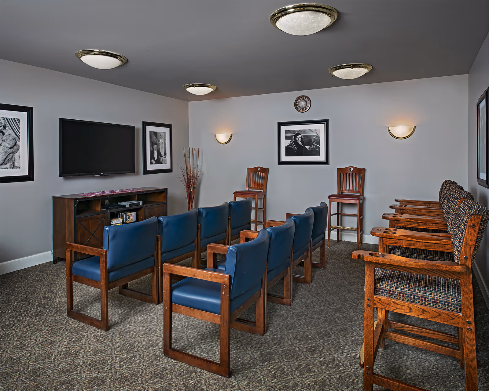 Small media room with rows of wooden chairs facing a wall-mounted TV and framed black-and-white photos on the walls.