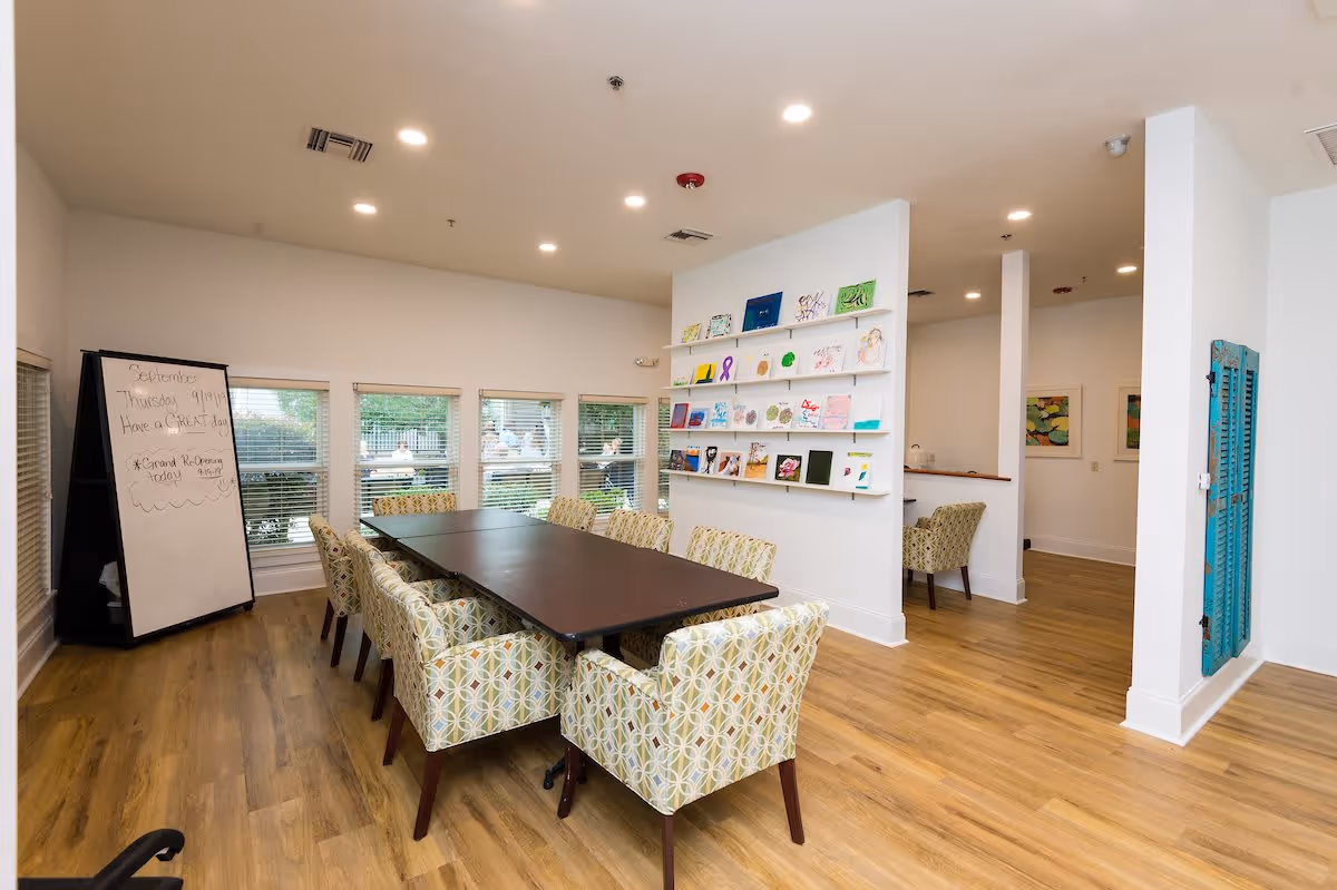 A bright and spacious interior room with a long dark wooden table surrounded by patterned upholstered chairs. There are large windows with blinds on one side, a whiteboard with writing on it, and a wall displaying colorful artwork. The room has light wood flooring and white walls, with recessed ceiling lights providing illumination.