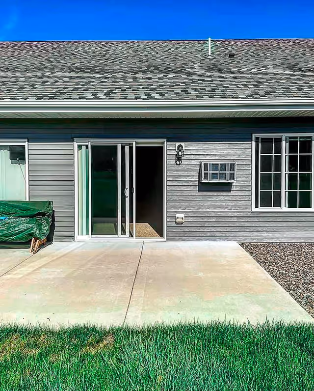 Exterior view of a single-story building with gray siding, a sliding glass door, a window with white trim, and a concrete patio in front. There is green grass in the foreground and a clear blue sky above.