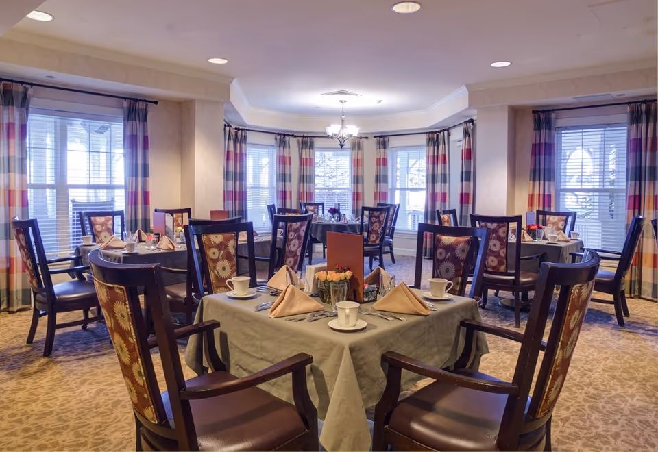 Bright dining room with multiple set tables, patterned chairs, and large windows with striped curtains.