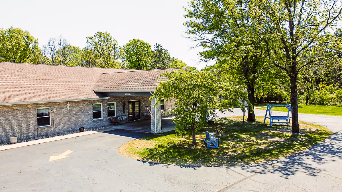 Exterior view of a single-story brick building with a covered entrance, surrounded by trees and greenery. There are two blue benches and a blue swing on a grassy area near the entrance, with a paved driveway and parking area in front.