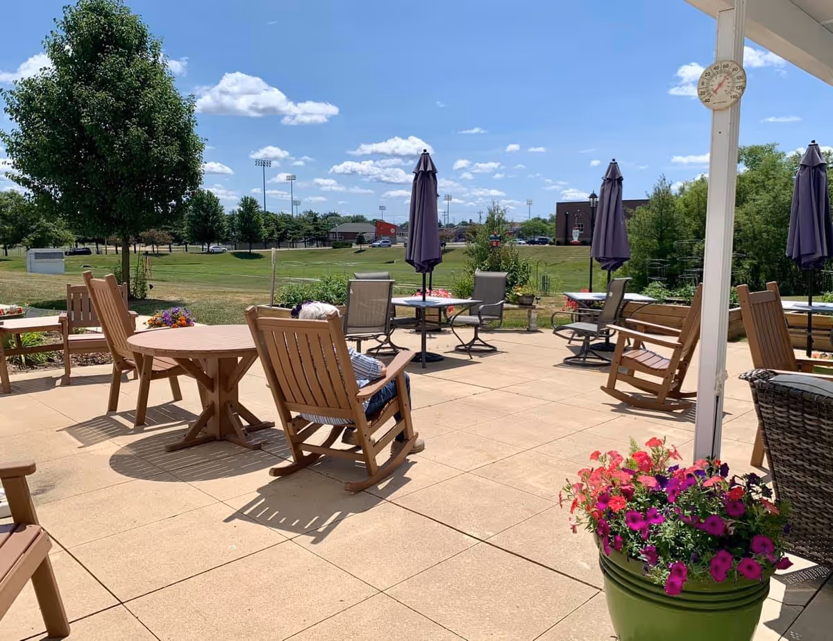 Outdoor patio area with several wooden rocking chairs and tables with closed umbrellas. A person is sitting in one of the rocking chairs. There are flower pots with colorful flowers and a green lawn with trees and buildings in the background under a blue sky with scattered clouds.