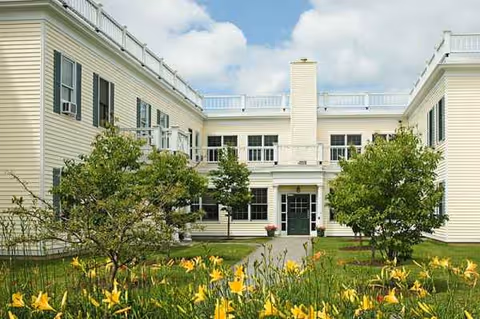 Cream-colored senior living center with a central entrance, surrounded by a garden and yellow flowers.