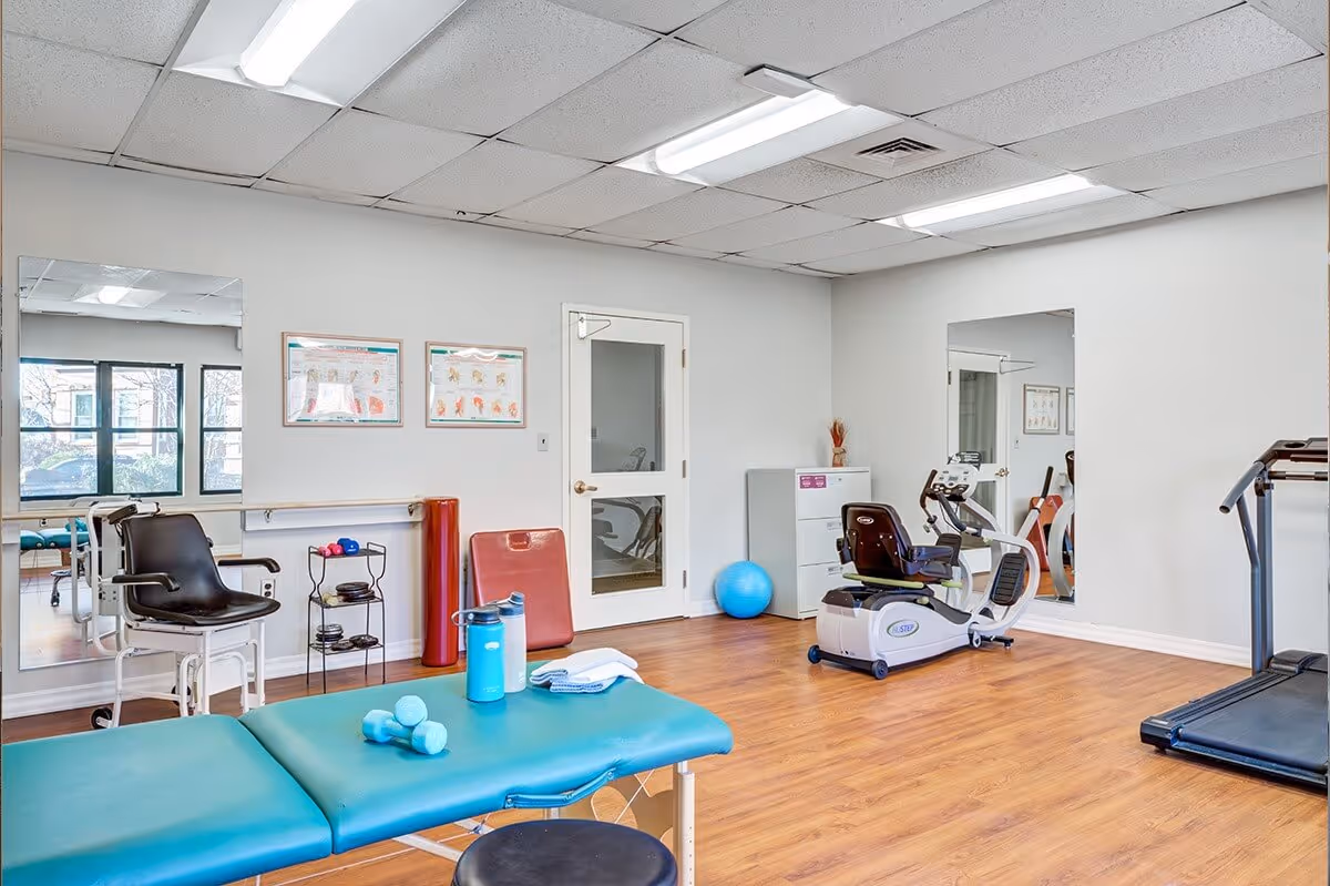 A bright physical therapy or exercise room with wooden flooring, featuring a teal therapy table with small dumbbells and a water bottle on it, an exercise bike, a treadmill, a blue exercise ball, and various exercise equipment. The room has white walls, large mirrors, and a window letting in natural light.