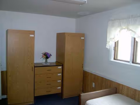 Senior living bedroom with wooden wardrobes, a dresser, and a bed by a window.