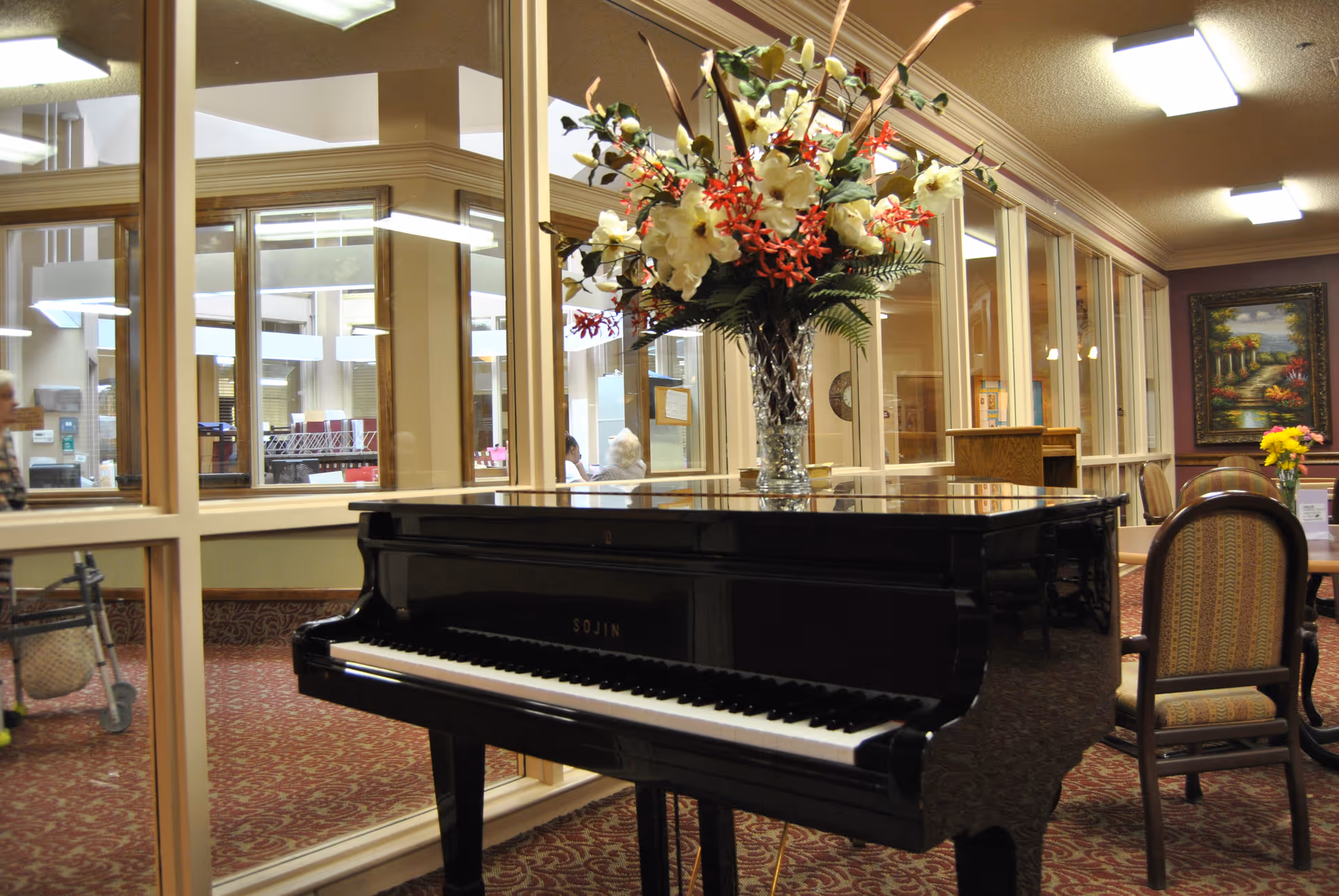 Interior view of a senior living facility common area featuring a black grand piano with a large floral arrangement on top. The room has carpeted floors with a patterned design, upholstered chairs around tables, and large windows separating this space from another area. There is a framed painting on the wall and ceiling lights providing illumination.