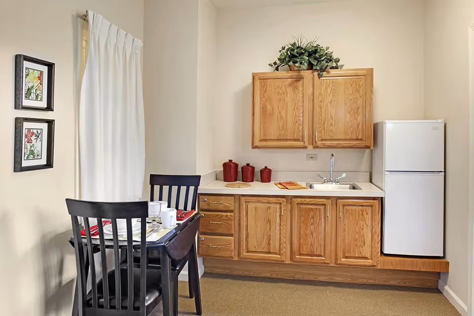 A small kitchen area with wooden cabinets, a white countertop, a sink, and a white refrigerator. To the left, there is a small dining table set for two with black chairs, white dishes, and red napkins. Two framed floral artworks hang on the wall next to a window with white curtains.
