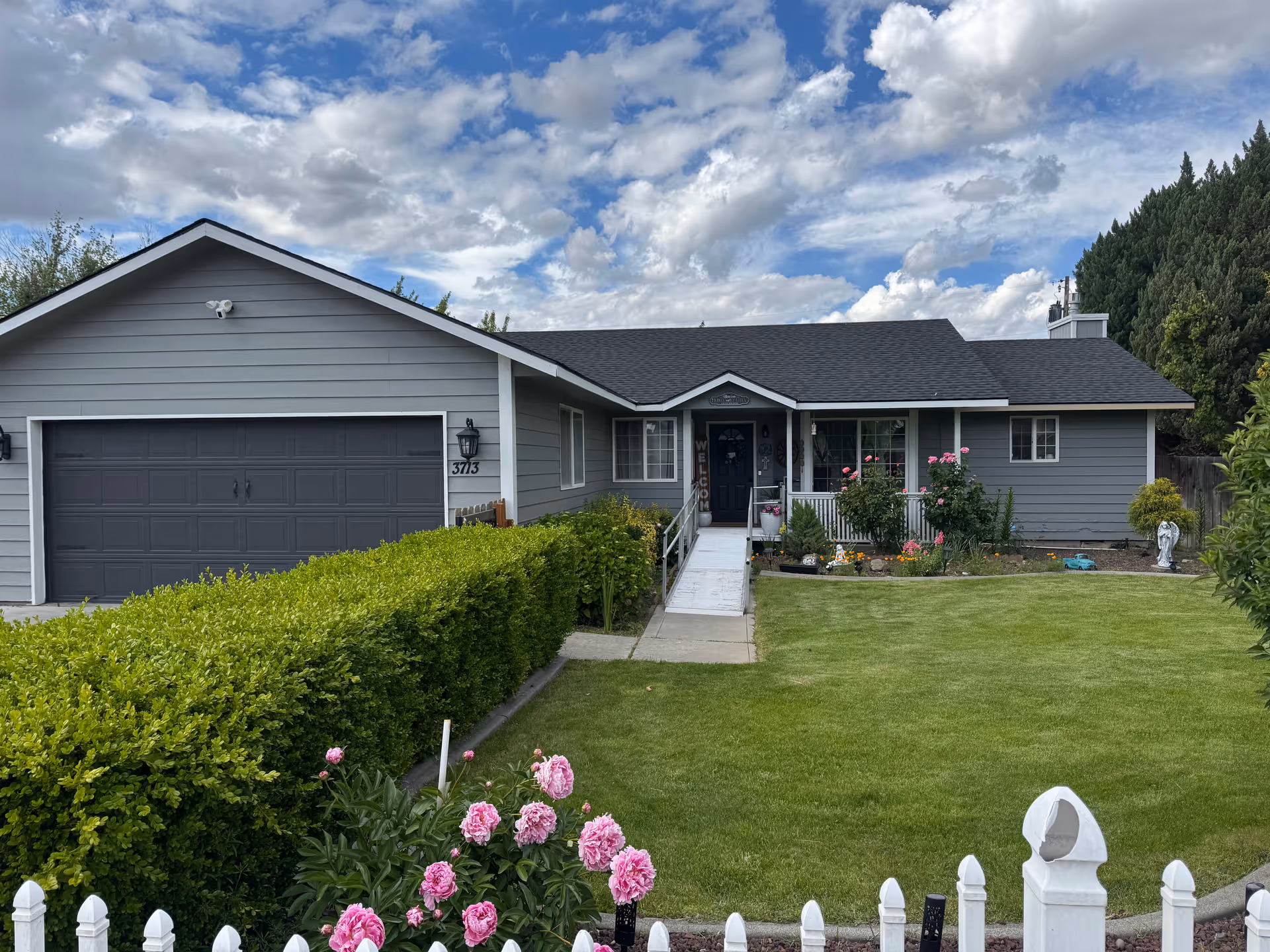 Front exterior of a gray single-story house with a garage, green lawn, flowerbeds, and a ramped walkway to the porch.