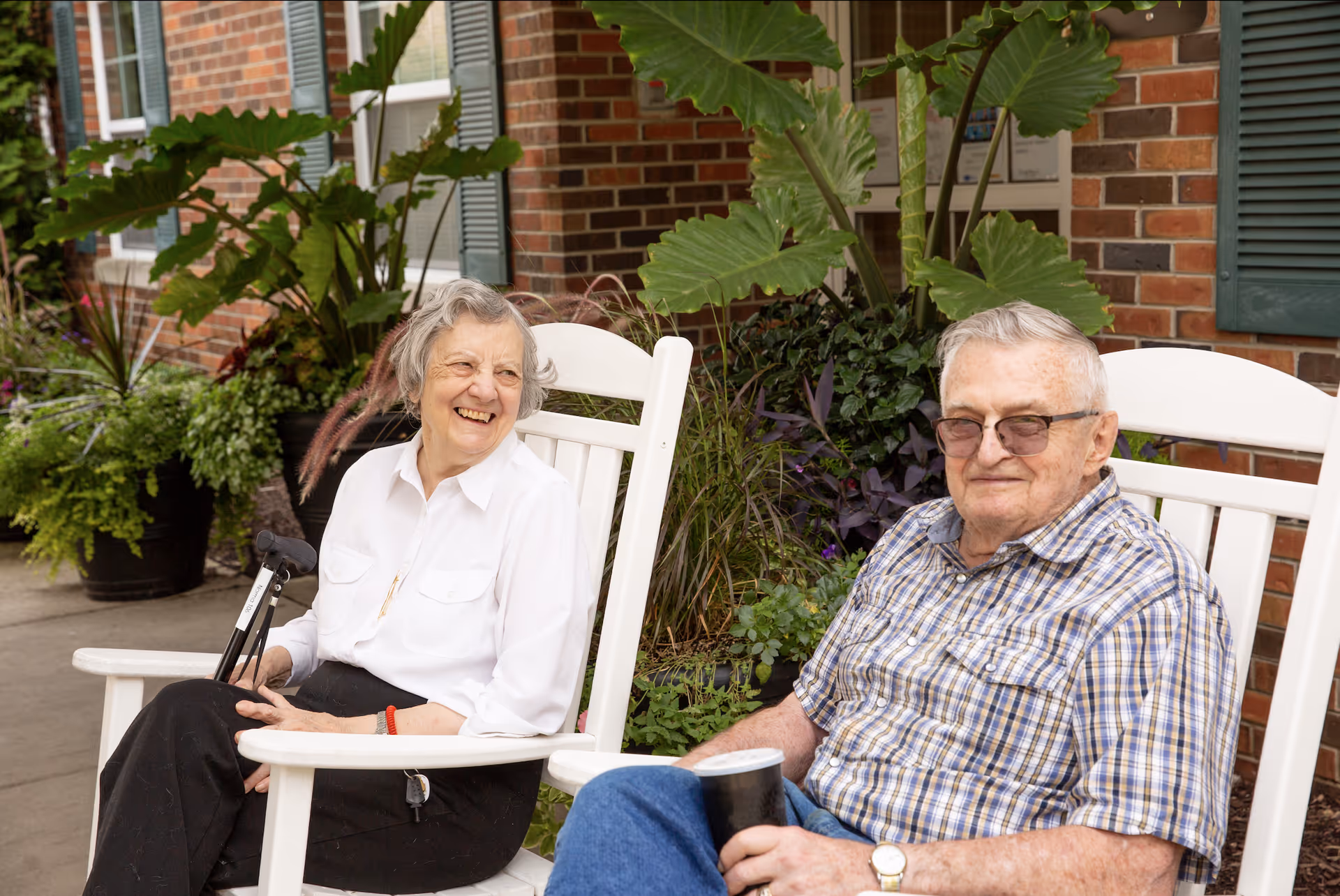 Two elderly individuals sitting and relaxing on white rocking chairs outside a brick building with green shutters and surrounded by potted plants and greenery. The woman is smiling and holding a cane, while the man is wearing glasses and holding a cup.
