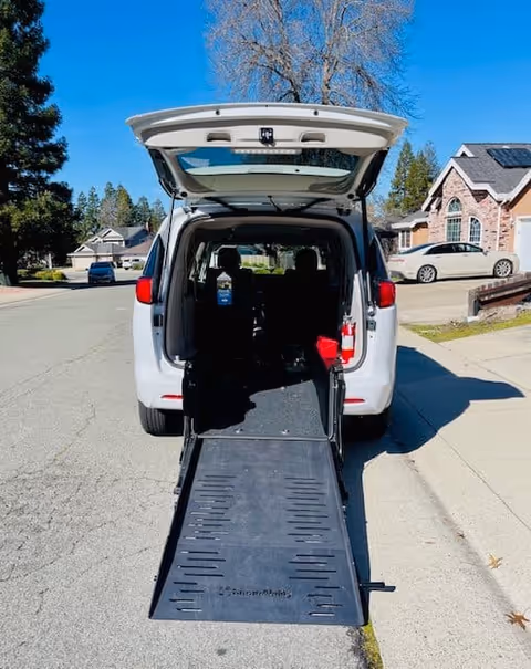 A white van with its rear door open and a wheelchair ramp extended onto a residential street with houses and trees in the background.