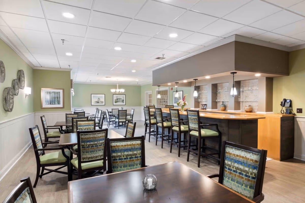 A well-lit dining area with multiple tables and chairs arranged neatly. There is a bar counter with high chairs on the right side, pendant lights hanging above it, and decorative items on the shelves behind the bar. The walls are painted light green with framed pictures and decorative wall hangings. The ceiling has recessed lighting and a chandelier is visible in the background.