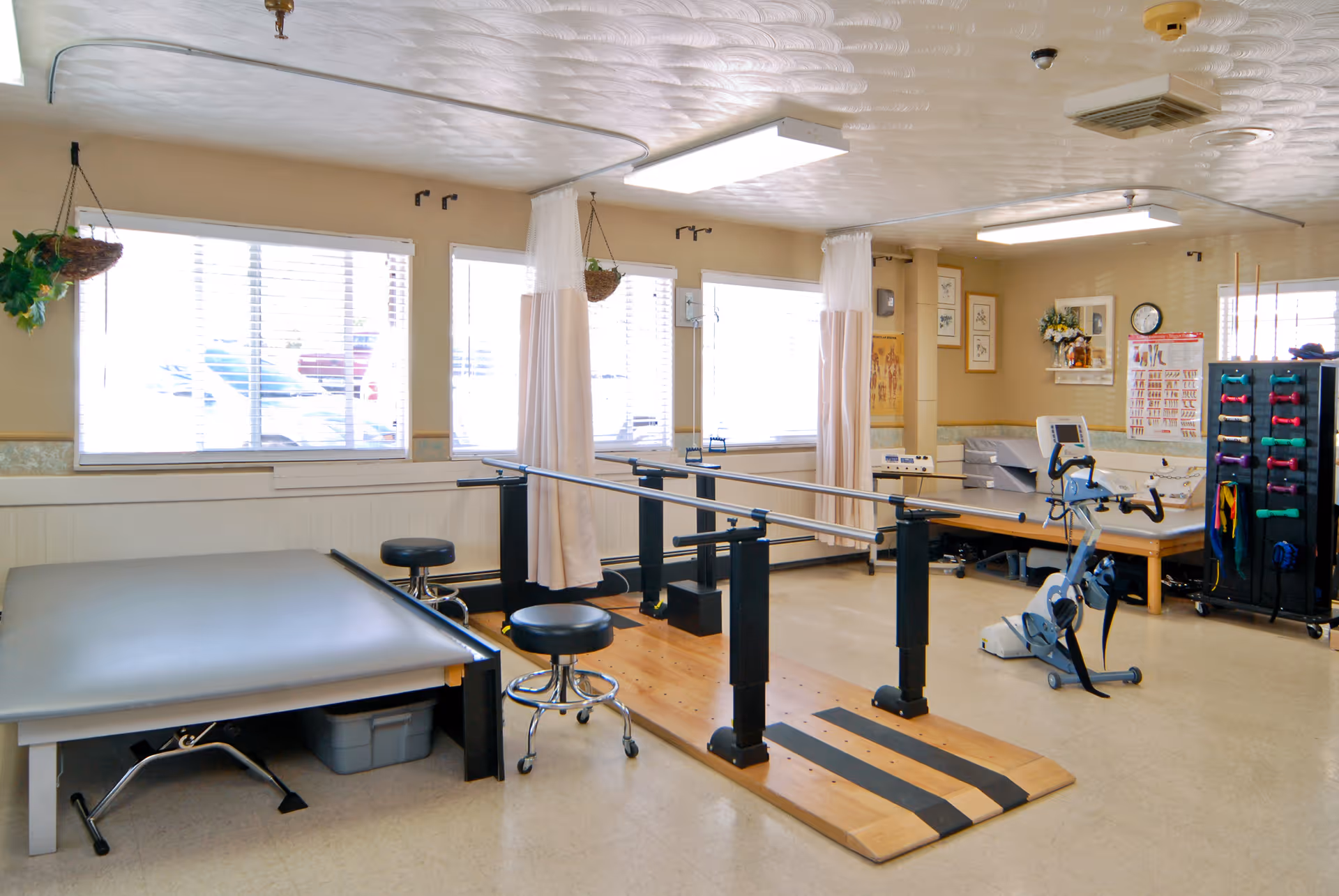 A physical therapy room with parallel bars for walking exercises, two black stools, an exercise bike, a padded treatment table, and a rack with colorful hand weights. The room has large windows with blinds, beige walls, and medical charts on the walls.