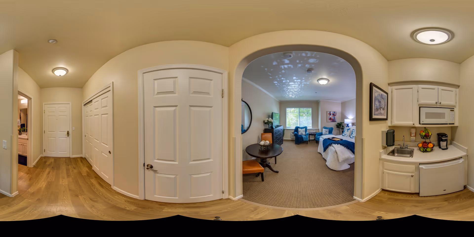 Interior view of a senior living facility apartment showing a hallway with wooden flooring, a small kitchenette with white cabinets, a microwave, sink, and coffee maker on the right, and a bedroom area with a bed, chairs, a round table, and a window in the background.