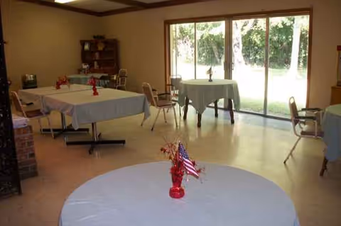 A dining room with round tables covered in blue cloths and red centerpieces by glass doors.