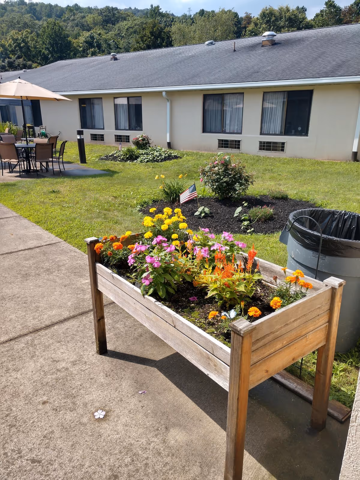 Outdoor garden area at Highland Manor featuring a raised wooden planter box filled with colorful flowers including pink, orange, and yellow blooms. In the background, there is a building with several windows, a patio area with tables, chairs, and an umbrella, a trash can, and a small flower bed with an American flag. Trees and greenery are visible beyond the building.