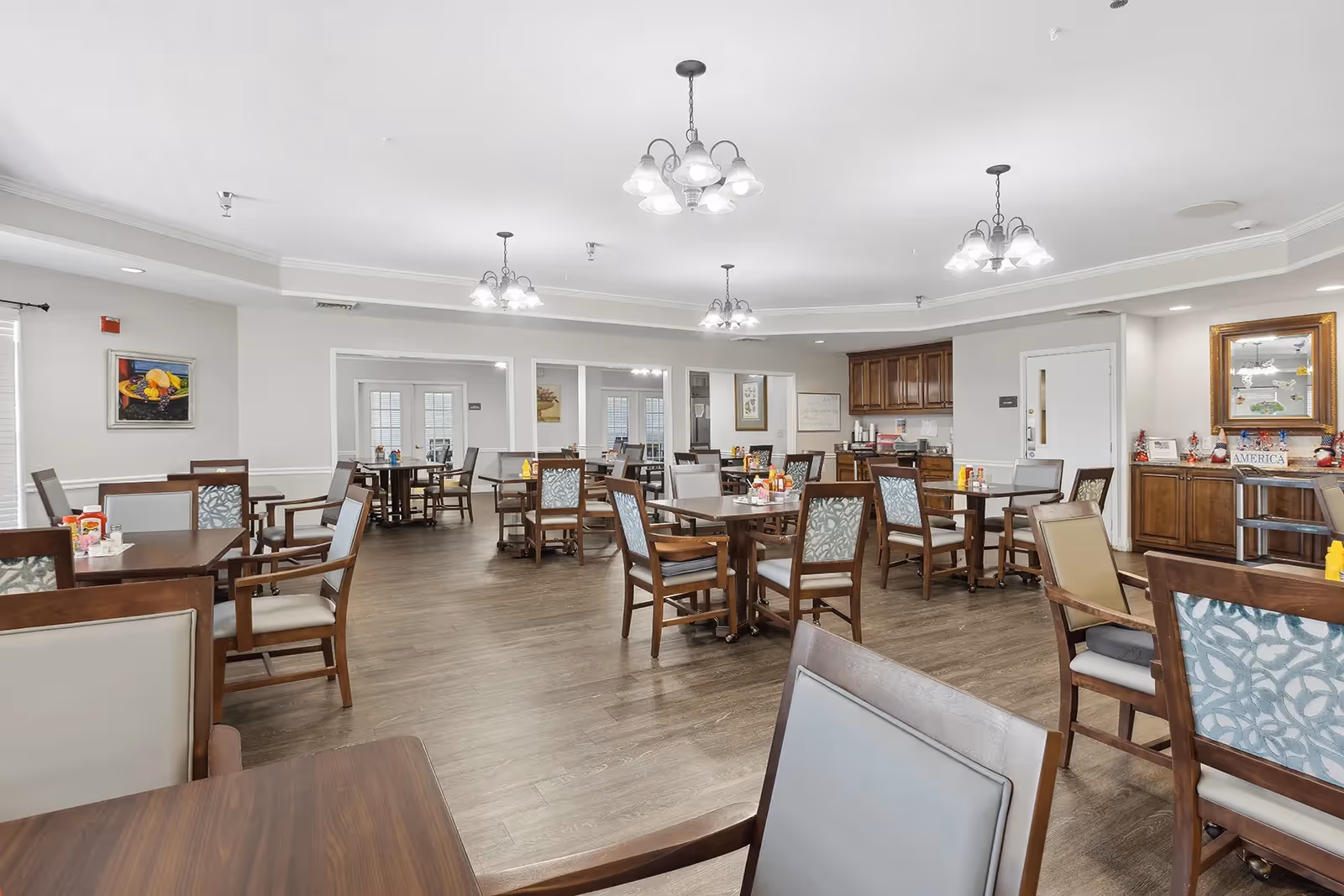 A spacious dining room with multiple wooden tables and chairs arranged neatly. The room has light-colored walls, several ceiling light fixtures, and wooden flooring. There are paintings and decorative items on the walls and cabinets, and a small counter area with coffee and condiments is visible in the background.
