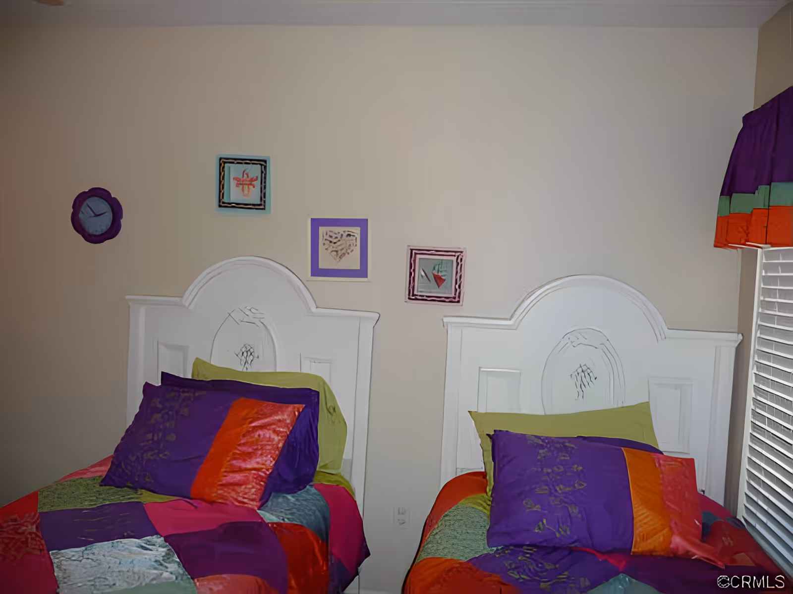 A bedroom with two twin beds featuring white wooden headboards. Each bed has colorful patchwork bedding in shades of purple, red, orange, green, and blue. Above the beds, there are three small framed artworks on the beige wall. A window with blinds and a multicolored valance is visible on the right side.