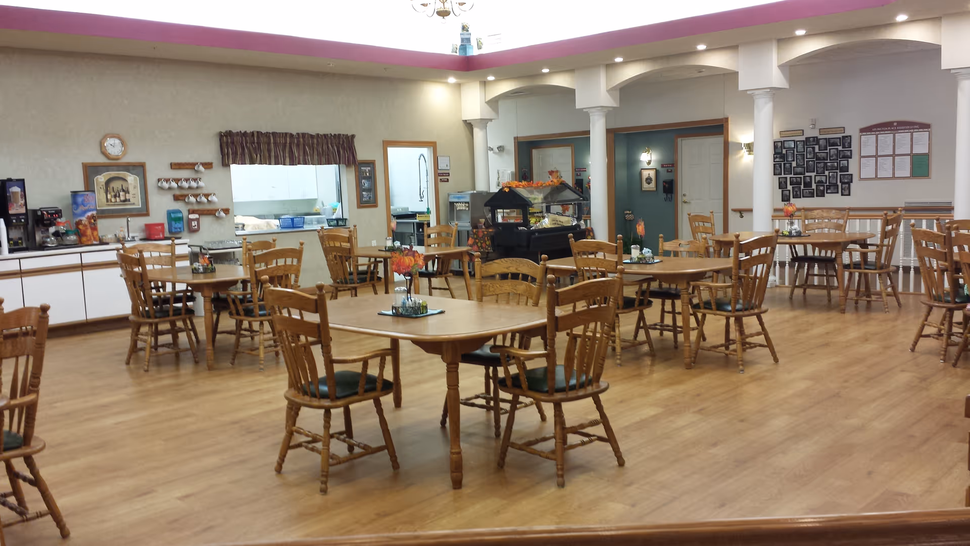 A spacious dining room with multiple wooden tables and chairs arranged neatly. Each table has a small floral centerpiece. The room has light-colored walls, a wooden floor, and a counter area with coffee machines and cups. There are columns and a decorative ceiling with recessed lighting.
